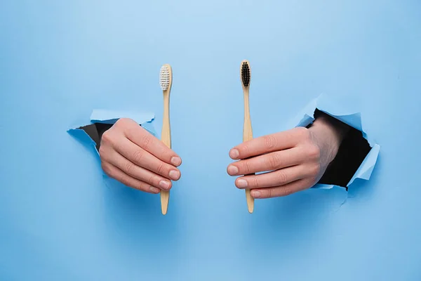 Unrecognizable female holding two bamboo eco toothbrushes through a torn blue paper wall. Dental care and Eco friendly and reuse concept. Copy space aside for your advertising content — Stock Photo, Image