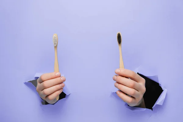 Unrecognizable female holding two bamboo eco toothbrushes through a torn purple paper wall. Dental care and Eco friendly and reuse concept. Copy space aside for your advertising content — Stock Photo, Image