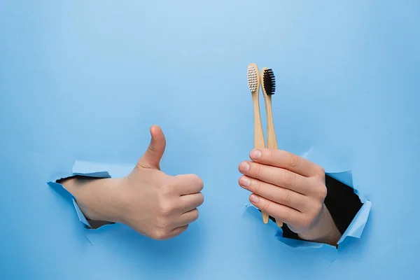 Unrecognizable female makes thumb up gesture, demonstrates bamboo eco toothbrushes, gestures through torn paper blue background. Body language concept. Dental care and Eco friendly and reuse concept — Stock Photo, Image