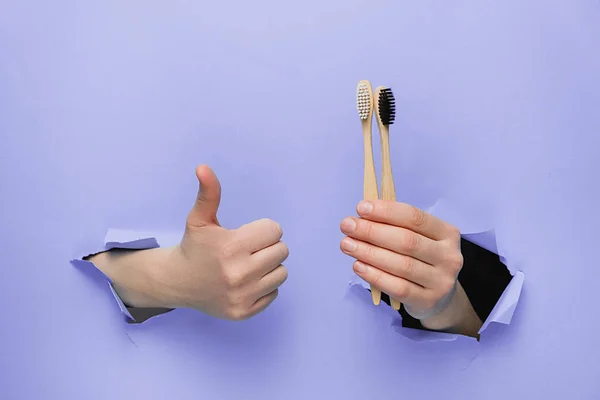 Unrecognizable female makes thumb up gesture, demonstrates bamboo eco toothbrushes, gestures through torn paper purple background. Body language concept. Dental care and Eco friendly and reuse concept — Stock Photo, Image