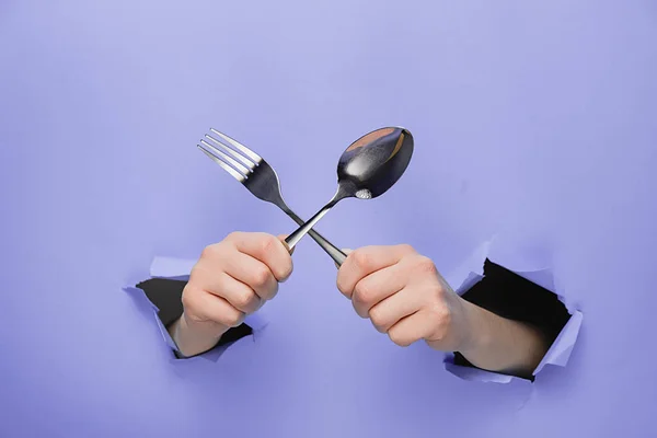 Female hands holding spoon and fork through torn purple paper wall. Gesture of eating dinning. Cooking, healthy homemade food concept. Copy space