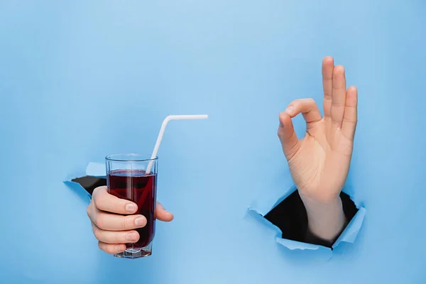 Close up of female hand holding a glass of fresh red juice with a straw through torn blue paper wall. Fruit drinks and healthy nutrition. Hand sign. Hole in wall. Like gesture — Stock Photo, Image
