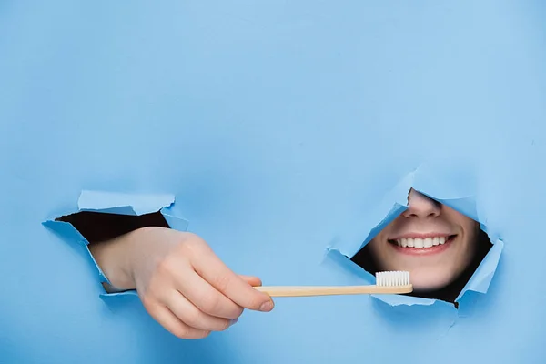 Woman with toothy pleasant smile on face, keeps head and bamboo eco toothbrush through torn hole in blue paper, copy space for your promotion. Dental care and Eco friendly and reuse concept — Stock Photo, Image