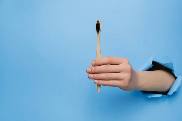 Close up of female hand holding bamboo eco toothbrush through torn blue paper wall. Dental care and Eco friendly and reuse concept. Copy space aside for your advertising content — Stock Photo, Image