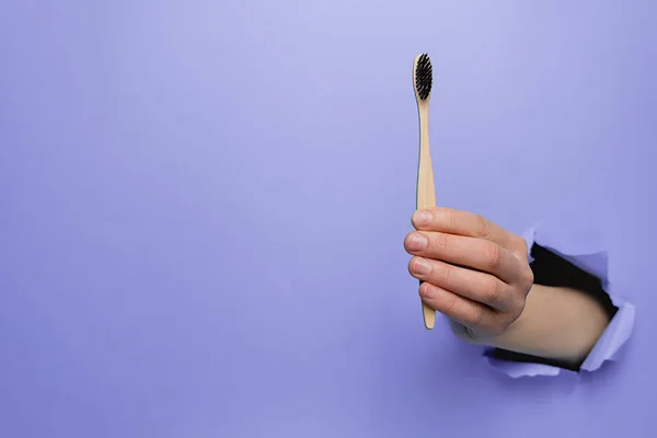 Close up of female hand holding bamboo eco toothbrush through torn purple paper wall. Dental care and Eco friendly and reuse concept. Copy space aside for your advertising content — Stock Photo, Image