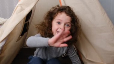 Little child vlogger saying hello hi looking at Camera at child room, cute kid talking to webcam making online recording having fun, curly girl with pretty face sitting in tent. slow motion