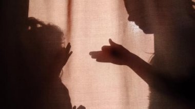 Silhouette of unrecognizable mother and child of primary school age telling stories using shadow presents talking dog's heads when sitting behind the curtains at home on a sunny day