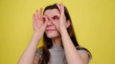 Funny young woman having fun making glasses shape with hands, does funny face with eyes wide open in studio isolated on yellow background, dressed in grey t-shirt. People emotions concept