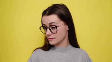 Dissatisfied woman imagining alternatives, weighs pros and cons, makes hard solution, spreads palms, cant choose answer, frowns face, wears spectacles and grey t-shirt, isolated over yellow background