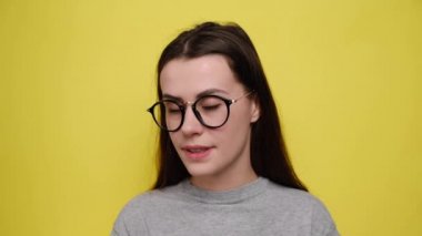 Wow! Happy young woman reacts on unexpected surprise, spreads palms raised up, grins happily at camera, dressed in grey t-shirt and eyeglasses, isolated over yellow background