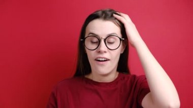 Şok olmuş, gözlüklü esmer kadın ellerini başının üstünde tutuyor, şaşırmış bir ifade takınıyor, ağzını iyice açıyor, tişört giyiyor, kırmızı arka planda poz veriyor. İnsan yüz ifadeleri. Tepki konsepti