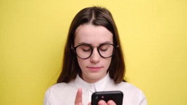 Şaşkın güzel bir kadın gözlüklü modern akıllı telefona bakıyor, şok edici haberler alıyor, ağzını kapatıyor, beyaz gömlek giyiyor, sarı arka planda izole edilmiş..