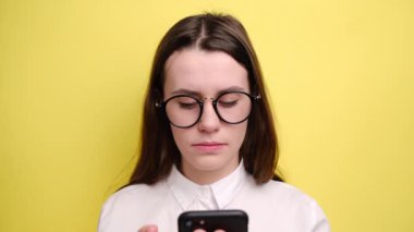 Gözlüklü mutlu esmer kadın modern cep telefonuyla internetten gelen harika davetiyeye bakıyor. Etkileyici iyi haberler okuyor, sarı arka planda izole edilmiş.