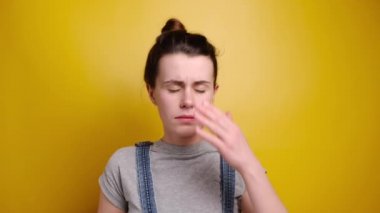 Frustrated woman holding hands on forehead being depressed regretting what she done having headache, wears grey t-shirt, stands over yellow background. Negative human emotions and feelings concept