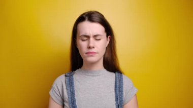 Hoşnut olmayan genç kadının boğazı ağrıyor, hasta ve sağlıksız, suratı asık, üşüttükten sonra acı çekiyor, gri tişört giyiyor, sarı arka planda duruyor. Rahatsızlık hastalıktan kaynaklanıyor.