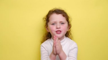 Yalvaran çocuk üzgün ve çaresiz görünüyor, af diliyor, üzgün ve suçlu hissediyor, avuç içlerini bir arada tutuyor, sarı arka planda duruyor. Sevimli şirin küçük kız bir şey için yalvarıyor.