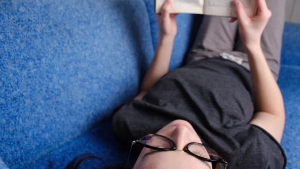 密切关注戴眼镜看旧书的女人 周末带着最喜欢的书在沙发上放松一下 在家里休息一天 松懈的年轻女子在客厅休息 — 图库视频影像