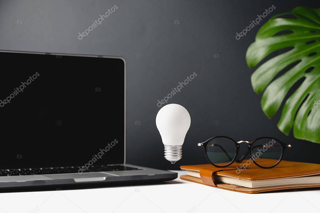 Home office desk table workspace with a notebook, light bulb, pen, cup coffee, green leaf monstera and eyeglasses on grey background. Copy space mockup template. Selective focus