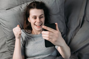 Uyanmış, heyecanlı genç bir kadının elinde akıllı telefon çığlığıyla yatağa uzanması ve sabahın ilk ışıklarıyla sevinç çığlıkları. Olumlu duygular ve mutluluk kavramı.