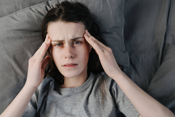 Несчастная задумчивая молодая женщина, держась за лоб, чувствуя себя одинокой, лежа в постели в одиночестве, пытаясь заснуть, думая о проблемах, страхах, заботах и безнадежности. Психологическая терапия, недостаток сна

