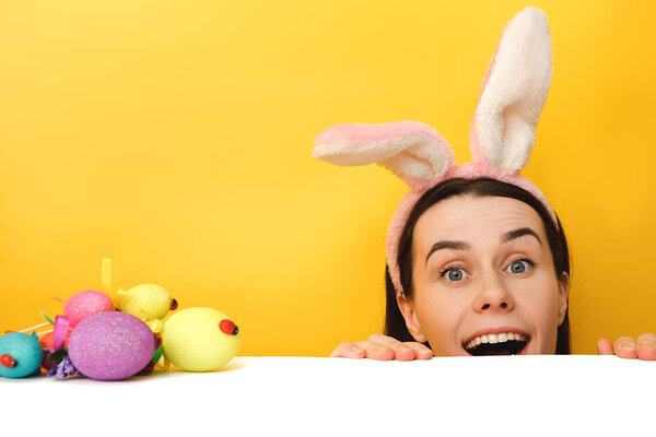 Studio shot of pretty woman sits at desk near colored Easter eggs, wears fluffy bunny ears, fun looks and hides face, models over yellow background with copy space. Spring holiday and Easter concept.