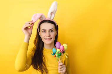Dost canlısı görünümlü mutlu kadının yakın çekimlerinde tüylü kulaklar takıyor renkli yumurtalar tutuyor, resim üstüne güzel bir ruh hali içinde, kazak giymiş, sarı arka planda soyutlanmış. Paskalya konsepti.