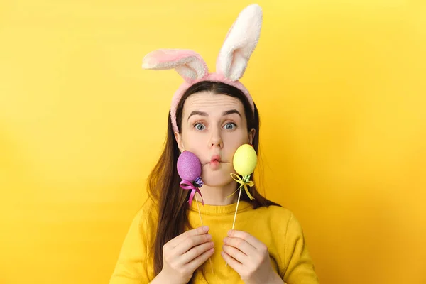 Comic young woman wears bunny ears makes funny face, plays fool, holds colored eggs, has happy expression, dressed in sweater, models over yellow background with copy space. Seasonal holiday concept