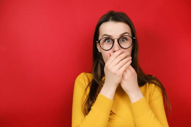 Gözlüklü ürkek kadın kameraya bakıyor, elleri ağızla kaplayan büyük gözleri dehşete düşürüyor, kırmızı stüdyo arka planında yalnız poz vererek şok edici haberler alıyor. Stresli durumlar kavramı