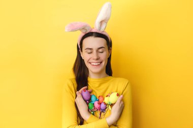 Güzel görünümlü, iyimser, pofuduk kulaklı, mutlu bakışlı, gözleri kapalı, boyanmış yumurtaları çubukların üzerinde tutan, sarı stüdyo duvarının üzerinde duran, metin için uygun yeri olan genç bir kadın..