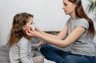 Anne, kızının evde oturup bulaşıcı hastalıklardan korunması için maske takmasına yardım ediyor. Anti-koronavirüs COVID-19 Salgın Hastalıkları Koruma Konsepti