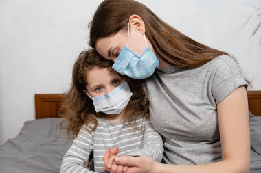 Evdeki yatakta oturan koruyucu maske takan genç bir anne ve küçük bir kızın portresi. Kendini izole et, ev karantinası. Koronavirüs veya COVID-19 salgın hastalık belirtileri