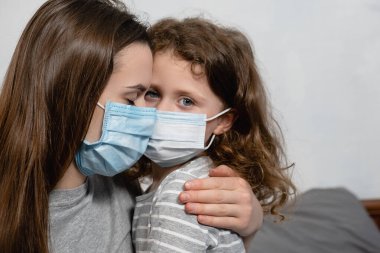 Virüs maskeli, üzgün, kıvırcık kızını kucaklayan, evde yatağında oturan, üzgün anaokulu kızını teselli eden annenin yakın portresi. Koronavirüs veya COVID-19 salgın hastalık belirtileri