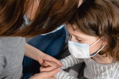 Yakın çekim üzgün, üzgün, yüzlü, maske takan, annesine sarılan, destek olan, sevgisini ifade eden küçük tatlı bir kız. Koronavirüs veya COVID-19 salgın hastalık belirtileri. Ev karantinası.