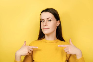 Genç bayan kendini işaret ediyor, kendi başarılarıyla gurur duyuyor, kameraya özgüvenli ve kendinden emin bakıyor, kazak giyiyor, sarı arka planda kopya alanı ile izole edilmiş. İnsanlar, gurur, kibir kavramı