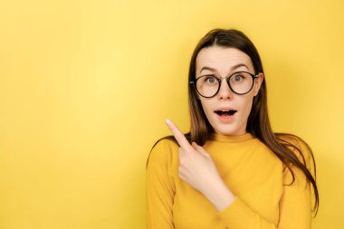 Gözlüklü güzel, neşeli bir kadının yatay görüntüsü ön parmağı uzakta, boş duvarda, sarı arkaplanda izole edilmiş bir kopya alanı gösteriyor..