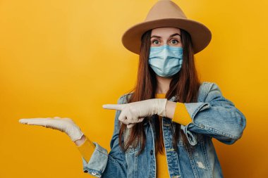 Shocked young woman in hat wears respirator mask to protect herself from coronavirus, demonstrates empty copy space for promo text, isolated over yellow background. Pandemic spreading virus concept clipart