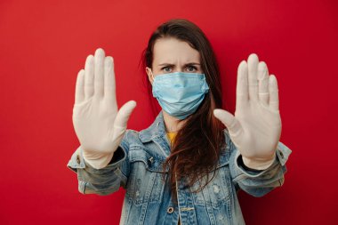 Steril maske takan ciddi bir kadın, kameraya doğru elini çekiyor dur hareketi yapıyor, limitini gösteriyor, kot ceket giyiyor, kırmızı arka planda izole edilmiş. Karantina salgını koronavirüs virüsü konsepti