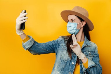 Elinde telefon tutan esmer kadın, sarı arka planda, kot ceket giymiş, kendini koronavirüsten korumak için koruyucu maske takan selfie çekiyor. Sosyal Mesafe kavramı