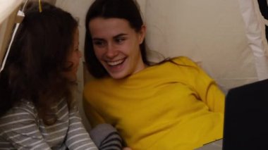 Gülümseyen genç anne ve küçük kız çocuğu evde çadırda dinlenen çizgi filmleri izlerken mutlu anne ve küçük kız komik videolar izliyorlar..
