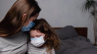 Endişeli annenin, virüs maskeli, üzgün, kıvırcık kızını kucakladığı, evde yatağında oturan, üzgün anaokulu kızını teselli ettiği portresi. Koronavirüs veya COVID-19 salgın hastalık belirtileri