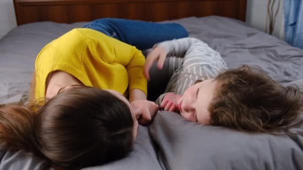 小女孩抱着吻着快乐的年轻妈妈躺在舒适的床上 笑着妈妈或保姆休息 女儿躺在床上和妈妈一起享受温馨的时光 亲属家庭概念 — 图库视频影像