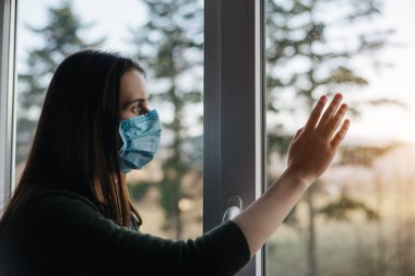 Tıbbi steril yüz maskesi takmış genç düşünceli bir kadın pencerenin pervazında oturuyor, camın üzerinde el ele tutuşuyor, bulanık pencereye bakıyor. Evde kalıp koronavirüs salgınını önleyin.