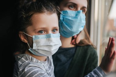 Mutsuz, genç bir annenin, virüs maskeli, üzgün, kıvırcık kızını kucaklaması, pencerenin kenarında oturup, üzgün bir anaokulu kızını teselli etmesi. Koronavirüs veya COVID-19 salgın hastalık belirtileri