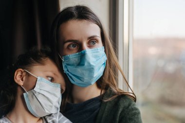 Tıbbi kısır maskeli, yorgun anne ve küçük kızı pencereye bakar, üzgün aile evde yalnız vakit geçirir. Koronavirüs veya COVID-19 salgın hastalık belirtileri