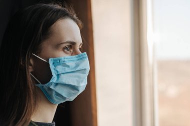 Koruyucu yüz maskesi takan genç bir kadın evdeki pencere pervazında oturuyor, bulanık pencereye bakıyor. Evde kalıp koronavirüs salgınını önleyin. Ev karantinası
