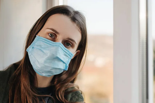 Close up portrait of brunette woman sit on windowsill with virus mask, thinks abot risk of epidemic disease, girl looks sadly at camera. Concept health and safety, N1H1 coronavirus, virus protection