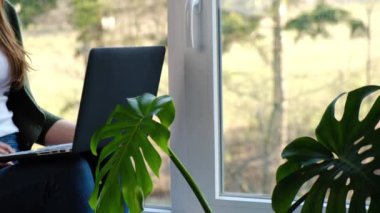 Modern dizüstü bilgisayarda çalışan ya da internetten bilgi öğrenen, pencere eşiğinde oturan, web kamerasıyla konuşan bir kızın videosu. Sosyal uzaklık