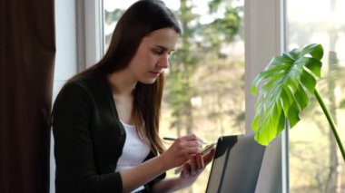 Düşünceli genç kadın Skype öğretmeniyle online çalışıyor ya da konferans dinliyor dizüstü bilgisayar başında oturup pencere eşiğinde oturup, notlar yazıyor, e-öğrenim eğitimi ve sosyal uzaklık kavramını anlatıyor.