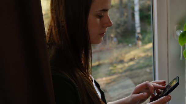 Vue de côté jeune femme assise sur le rebord de la fenêtre à la maison tenant le téléphone dans les mains textos message ou à l'aide d'applications mobiles, vérifier les applications de médias sociaux sur smartphone. Distance sociale. Mouvement lent 