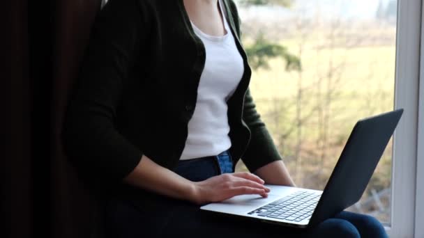 年轻女人在家里的窗台上用笔记本电脑 黑发女孩在社交网络上看新闻 在电脑上工作 在网上写博客或上网购物 概念上的社会疏远 — 图库视频影像